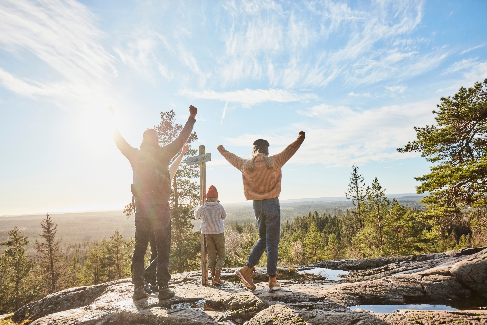 Familj som står med händena upp i luften på en utsiksplats i skogen