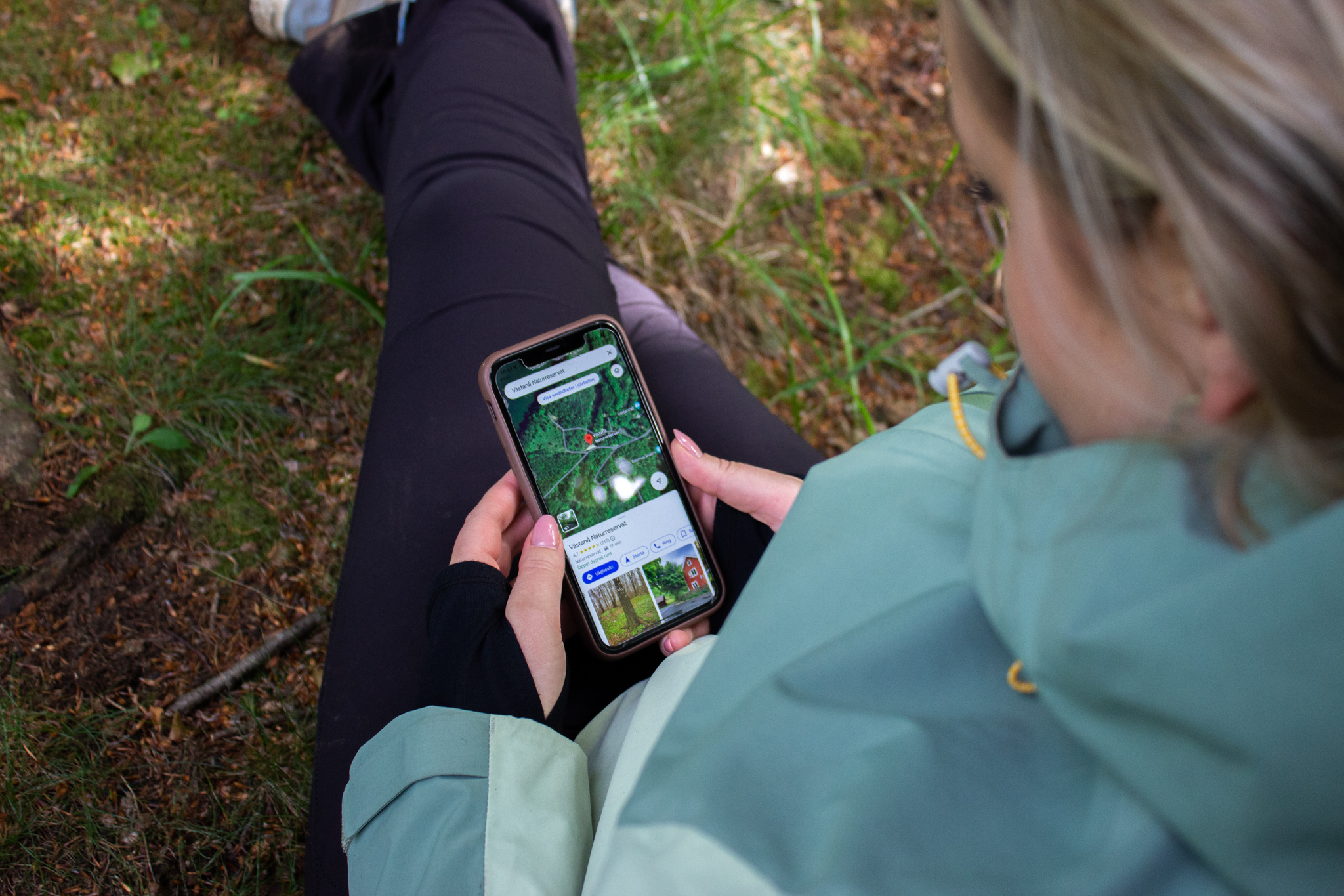 Kvinna som håller fram sin mobil och tittar på en Googlse maps sida medans hon sitter mot en tall i skogen.