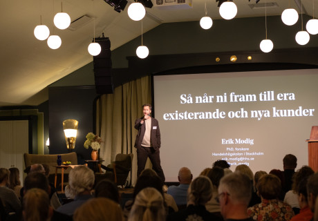 Erik Modig som står på scenen och föreläser om hur du når ut till nya och befinliga kunder. Publik runtomkring, slides bakom honom, bollformade lampor som hänger över publiken.